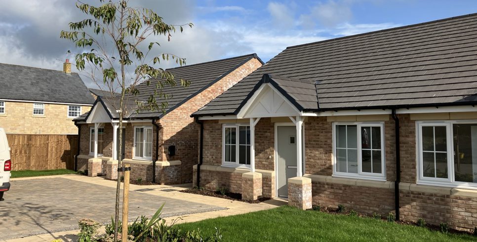 two newly built bungalows with light bricks and slate roofs. They have a small garden and driveway.