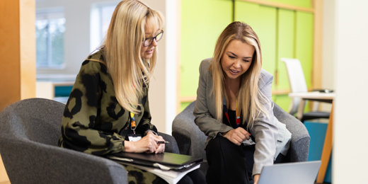 Two people sat next to other other at a table looking and pointing at a laptop. The setting is an informal meeting in a bright office.