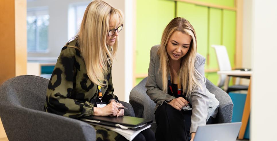 Two people sat next to other other at a table looking and pointing at a laptop. The setting is an informal meeting in a bright office.