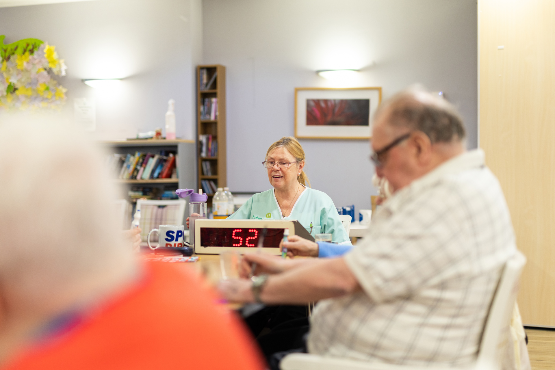 A person in a carer's uniform leads a game of bingo for a group of people sat at a table. 