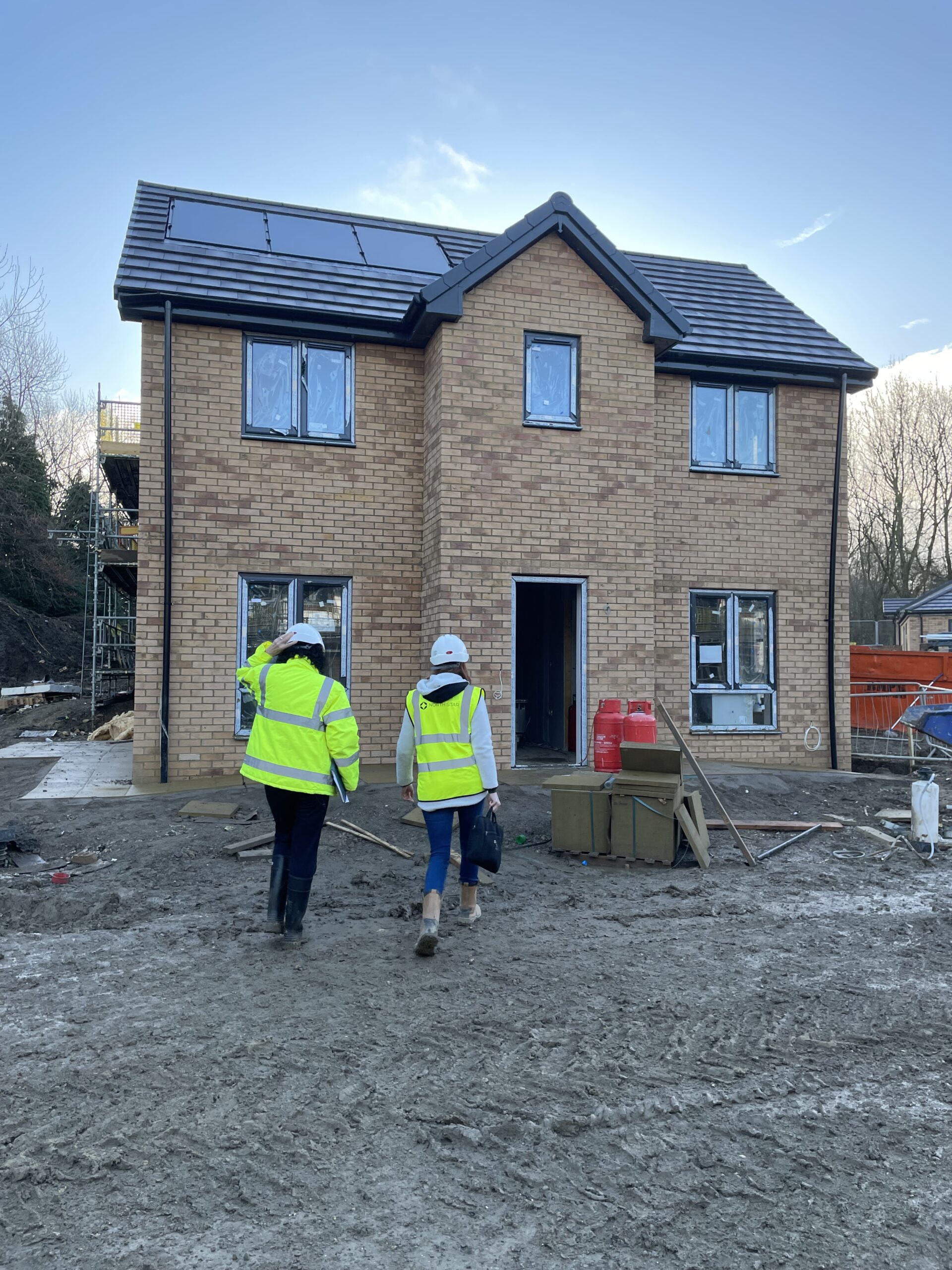 Two people wearing high-visibility jackets and hard hats walk toward a newly constructed brick house on a muddy building site. The house has solar panels on the roof and windows covered in protective film. Construction materials and equipment are scattered around the site.
