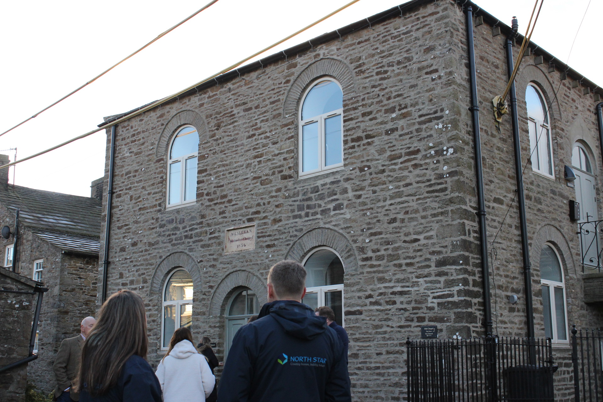 A group of people, including someone wearing a North Star Housing Group jacket, stand outside a renovated stone building with arched windows. The scene shows part of an opening event for the converted chapel, which now contains two flats, taking place in rural Bainbridge.