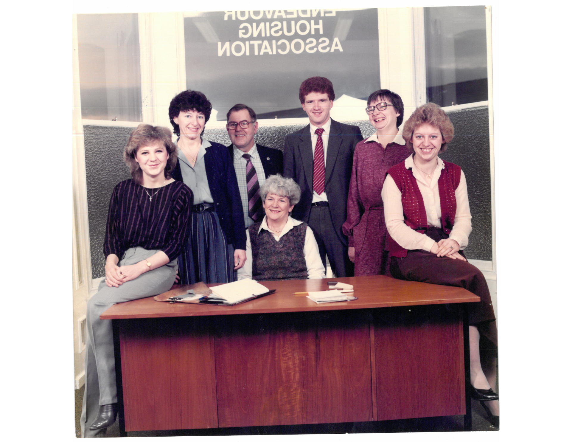 A vintage photograph of eight people posing together in an office setting, with "Endeavour Housing Association" visible in reverse on the window behind them. Two women are seated on a wooden desk at the front, while the others stand behind it, smiling. The group appears to be staff members from an earlier era, dressed in formal and semi-formal clothing typical of the late 20th century.