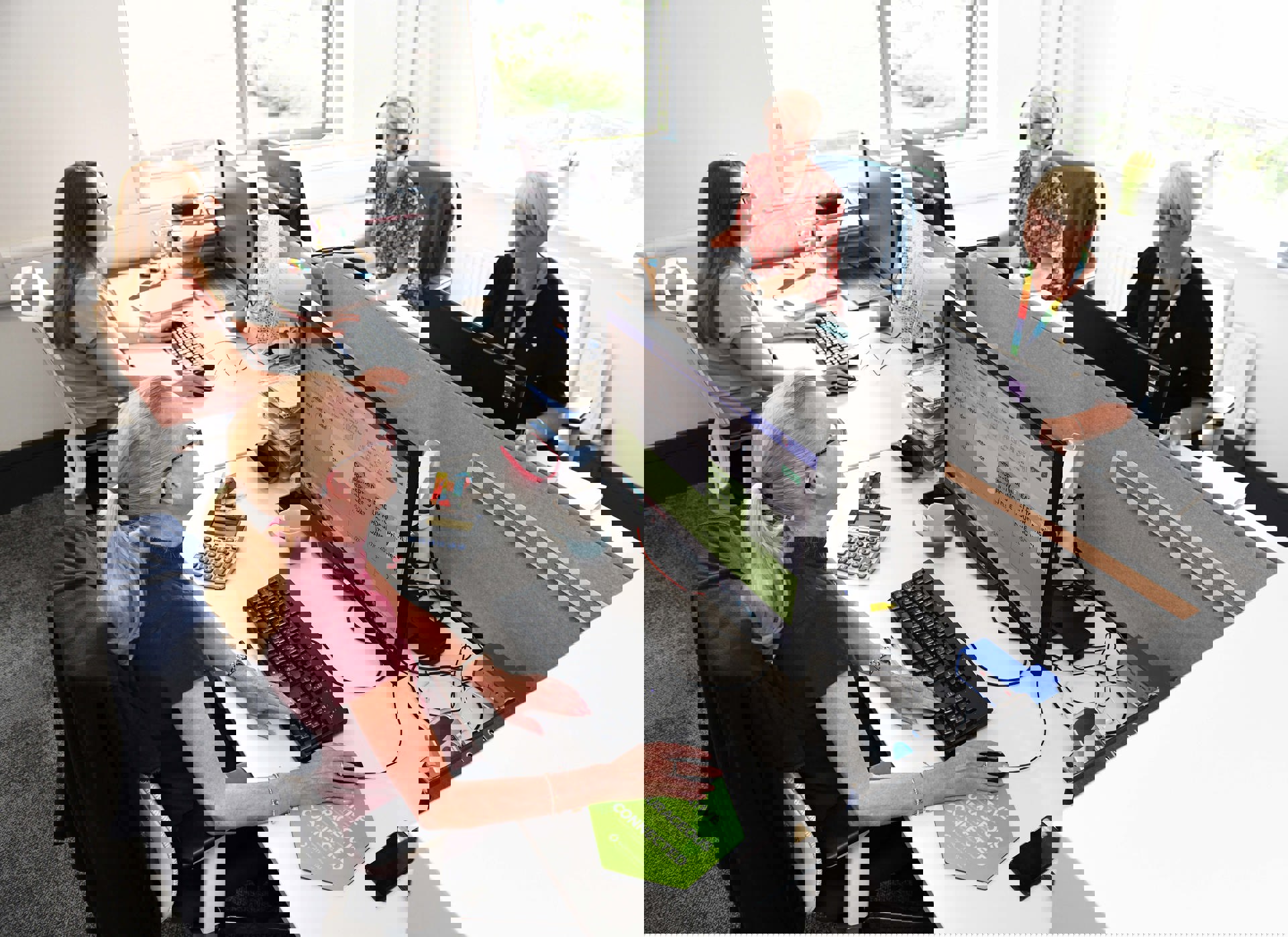 Four women are working at their desks in a bright, modern office. They are using desktop computers and appear focused and engaged. One woman is wearing a headset, and others are smiling and interacting. Office supplies, mugs, and documents are neatly arranged on the desks. A green sign on one desk displays the words "Creative, Courageous, Connected," reflecting North Star's values.