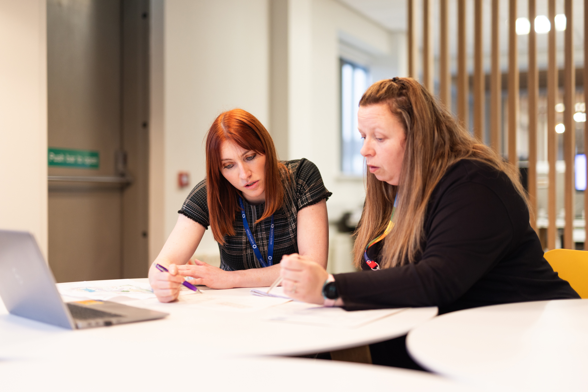 Two women seated at a round white table in a modern office space, collaborating on documents. One woman with red hair in a checked dress is attentively reviewing papers, while the other woman with long brown hair and a black top gestures towards the documents. Both are wearing lanyards, and a laptop is open on the table beside them. The background features vertical wooden slats and large windows.