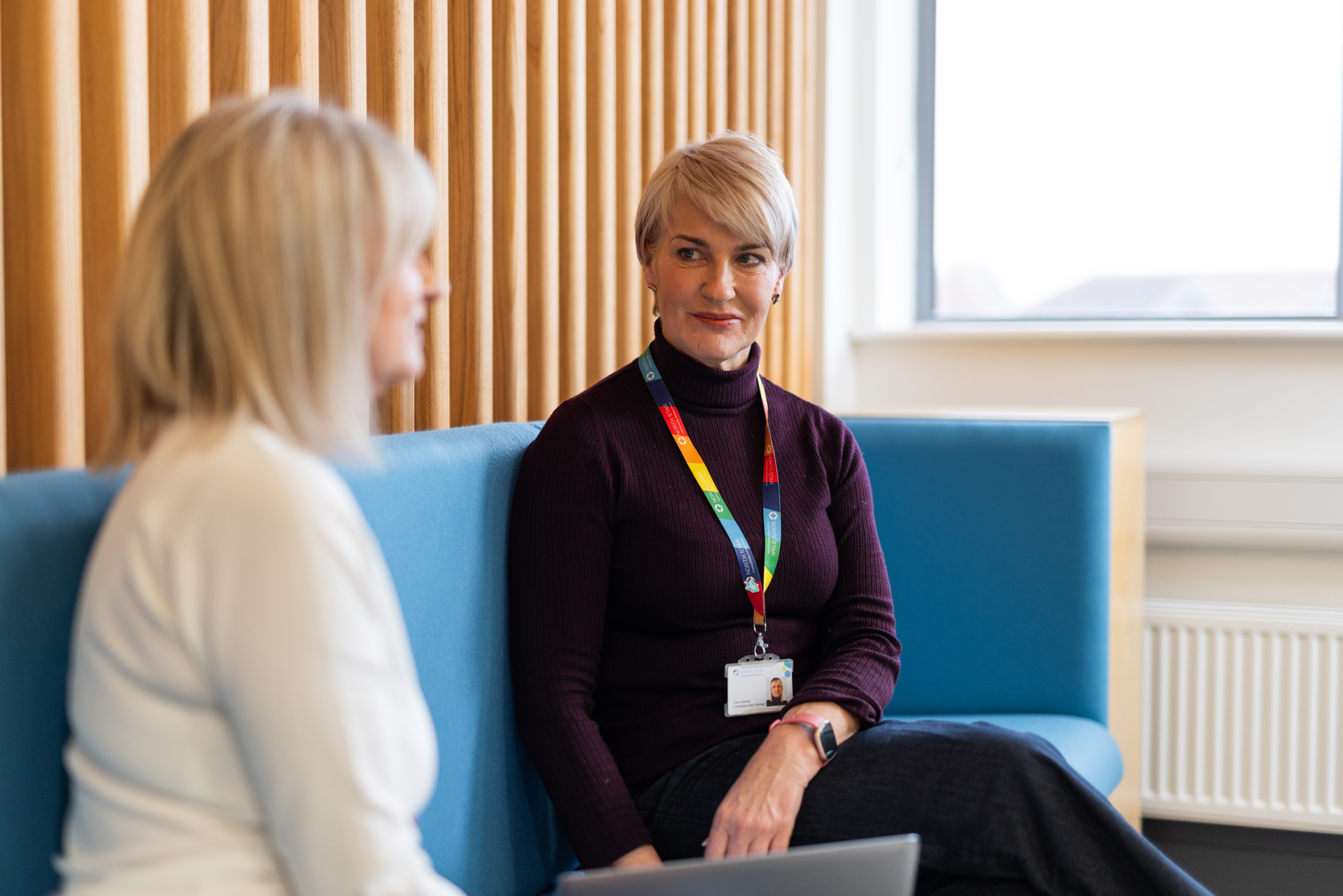 A person looks towards someone sat next to them with a pleasant expression on their face. They have a laptop in front of them and are sat in a communal space in an office.