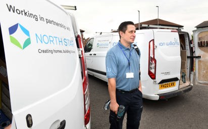 A repair worker stands between two branded vans from Ian Williams and North Star Housing. He is holding tools and wearing a blue uniform and lanyard. The vans display the logos and partnership between Ian Williams and North Star Housing.