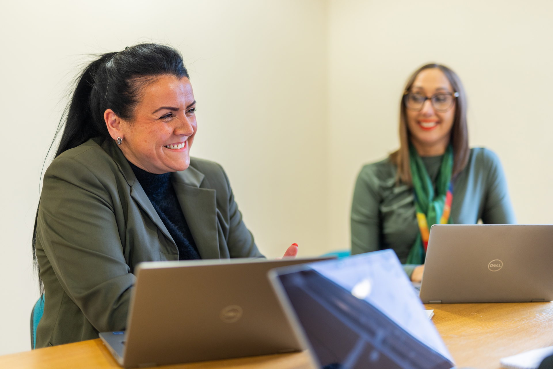 Two people sat next to other other at a table with laptops. The person closest to the camera is in focus and is smiling joyfully.