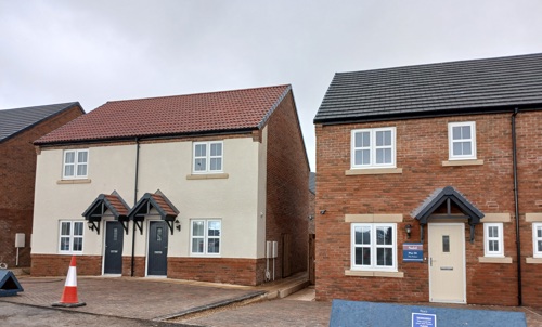 Two semi-detached homes with a freshly paved driveway. The building on the left has a red roof, black front doors with a canopy over each and a cream facade. The building on the right has a black roof, red brick facade and cream front door with canopy..