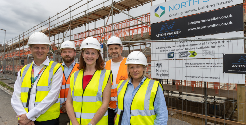 A group of people wearing hard hats and high vis vests stand outside an active building site smiling