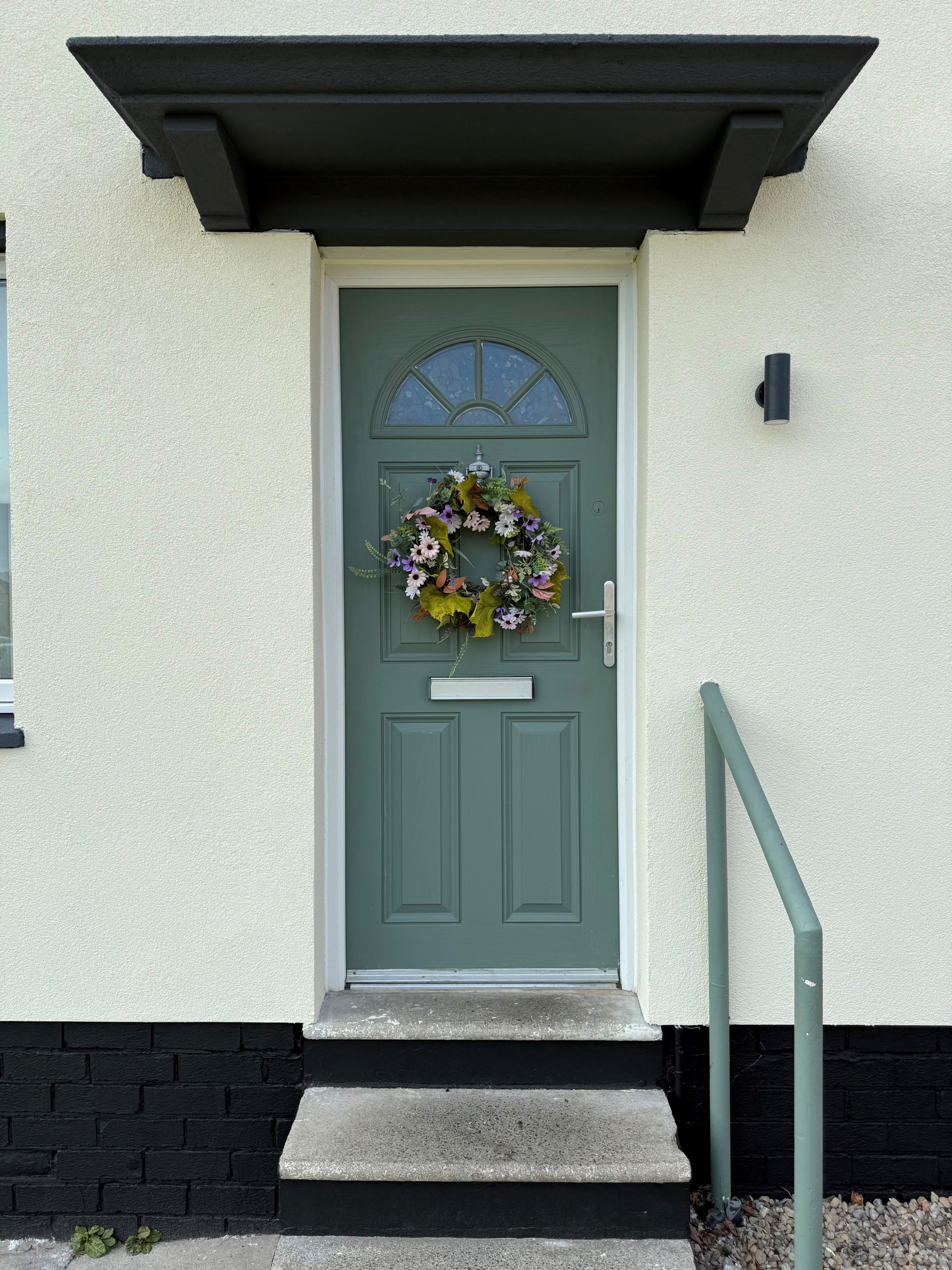 A green front door with two steps leading up to it, a hand rail for assistance and a flowery wreath.