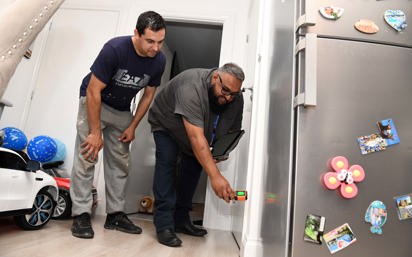 A housing officer uses a handheld device to inspect a wall inside a home, while the resident watches closely. They are in a kitchen area with a fridge covered in magnets and children’s toys visible in the background.  Let me know if you want a version tailored for accessibility compliance or a shorter version for web use.