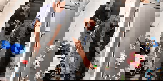 A housing officer uses a handheld device to inspect a wall inside a home, while the resident watches closely. They are in a kitchen area with a fridge covered in magnets and children’s toys visible in the background.  Let me know if you want a version tailored for accessibility compliance or a shorter version for web use.