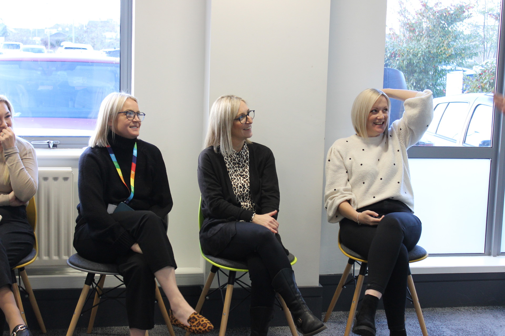 Four women sitting on chairs in a bright room, smiling and engaged in conversation. They are dressed in smart-casual clothing, with two of them wearing glasses. One woman has a rainbow lanyard, and another is casually resting her arm on her head. Large windows behind them reveal parked cars and greenery outside