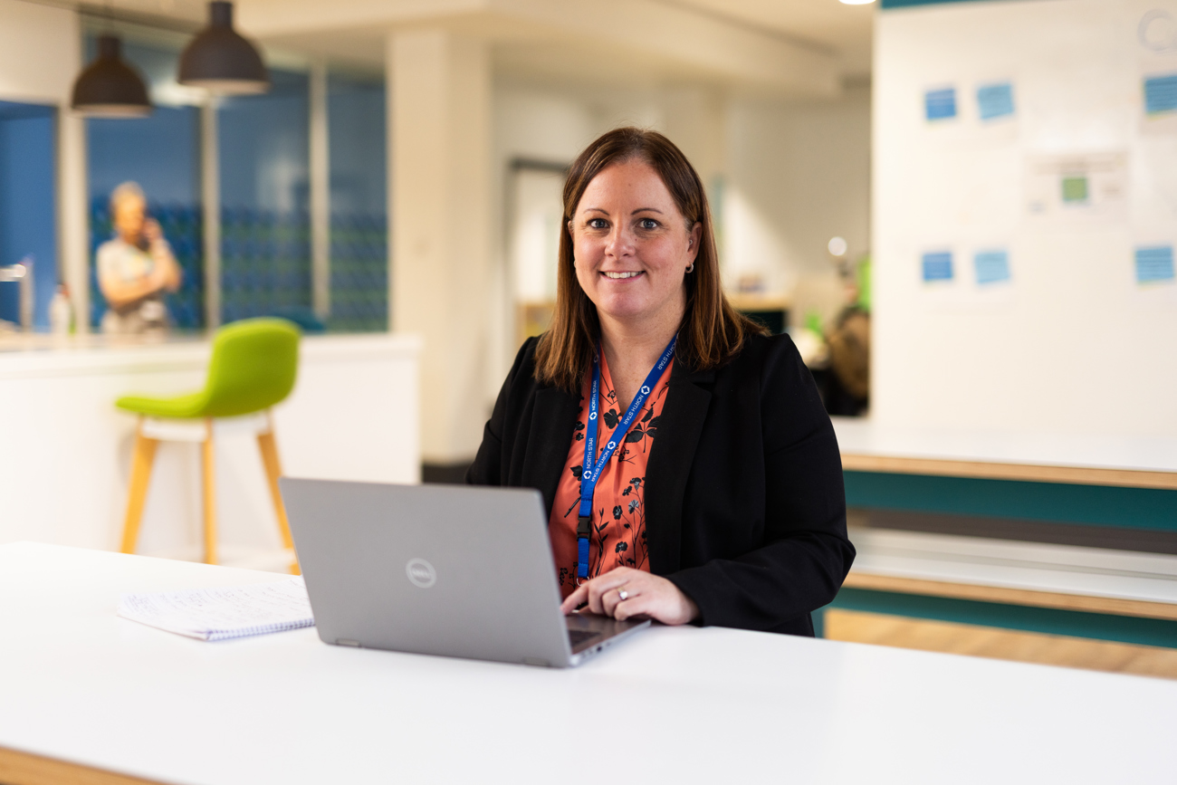 Woman smiling while working on a laptop at a bright office desk, wearing a black blazer and an ID badge.