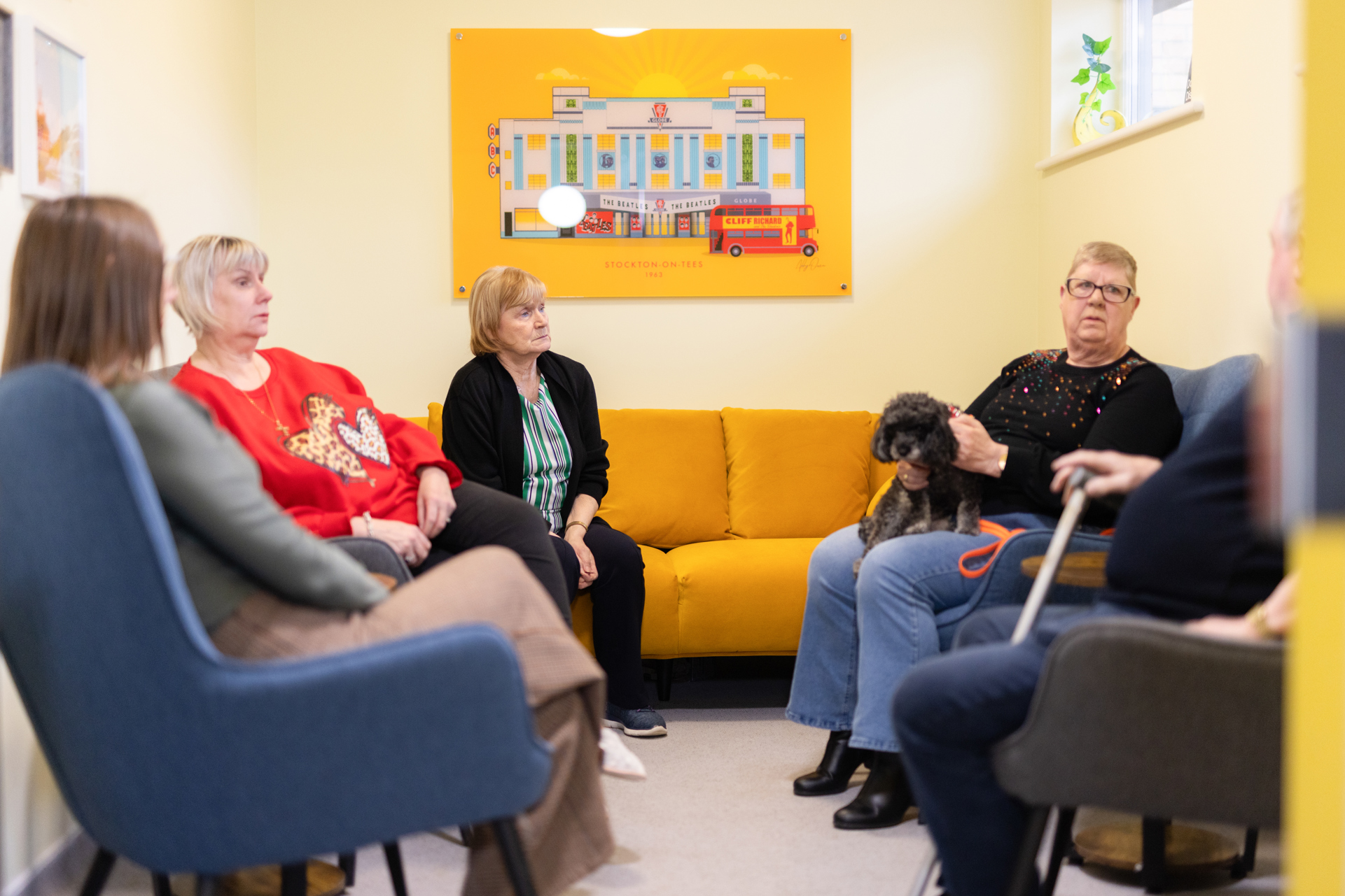 A group of people and a dog sat talking in a brightly lit and decorated communal space.