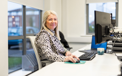 A woman with long blonde hair sits at a desk in an office, smiling at the camera. She is wearing a patterned black and white blouse and using a computer mouse. A keyboard, monitor, and a blue and white mug are on the desk in front of her. Large windows in the background show a car park and buildings outside