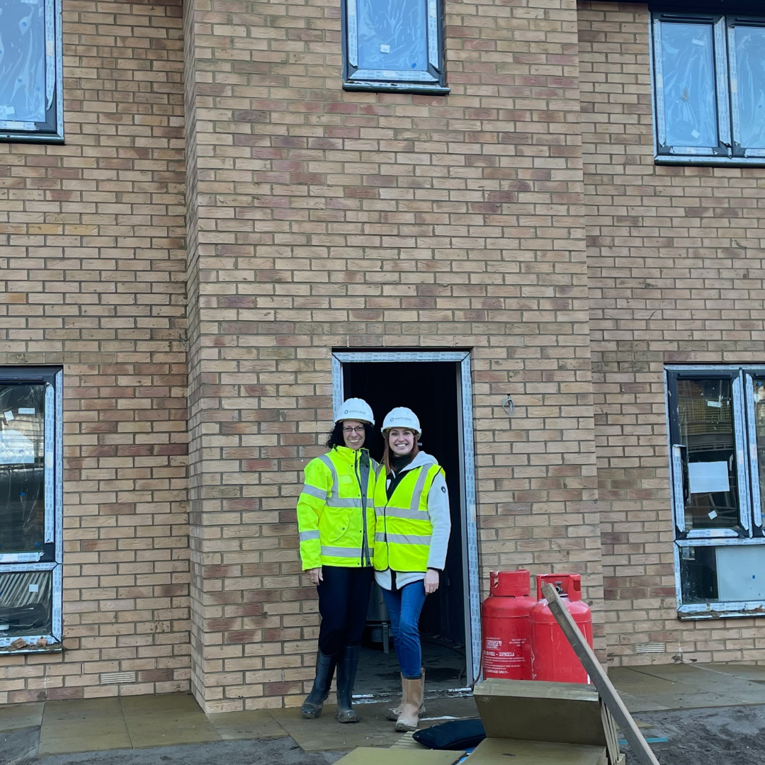 Two women smiling wearing hard hats and high vis vests stand in front of a home in development. The building has light coloured bricks and there are signs that this is a construction site such as gas canisters, muddy ground, and stickers over the window frames.