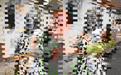 A smiling woman sitting in a wheelchair outside her home, holding a fluffy white dog on her lap and a large mug that reads 'Magnificent Mum.' She is wearing a white dress with green and black spots. Behind her is a white front door with a floral wreath and a garden with colorful potted flowers.