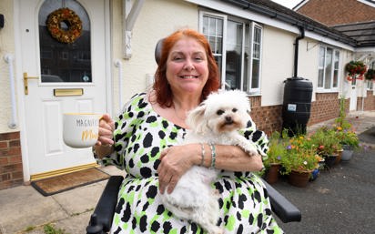 A smiling woman sitting in a wheelchair outside her home, holding a fluffy white dog on her lap and a large mug that reads 'Magnificent Mum.' She is wearing a white dress with green and black spots. Behind her is a white front door with a floral wreath and a garden with colorful potted flowers.